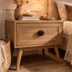 Könnte beinhalten: Ein hellbrauner Nachttisch aus Holz mit einer Schublade und einem dekorativen Knopf. Der Nachttisch hat konische Beine und ist mit einer Vase mit Trockenblumen, Büchern und einer Kerze ausgestattet. Ein geflochtener Korb und ein Teppich befinden sich im Vordergrund.