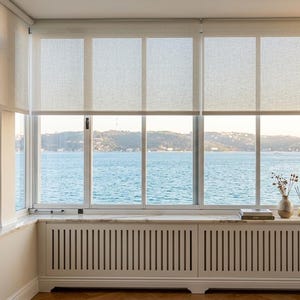 Op de afbeelding: Een grote raampartij met witte kozijnen en semi-transparante rolgordijnen. Onder het raam bevindt zich een witte radiatorombouw met verticale lamellen, afgewerkt met een marmeren vensterbank waarop een vaas en kleine decoratie staan.