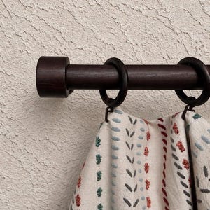 Puede incluir: Una barra de cortina de madera de color marrón oscuro con anillas a juego que sostiene una tela de color crema con filas verticales de puntadas bordadas en tonos azul, rojo y verde. La barra tiene un remate cilíndrico a la izquierda.