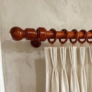 Può includere: Un bastone per tende in legno marrone scuro con un'estremità decorativa e anelli. Il bastone sostiene tende plissettate bianche. L'hardware è montato su una parete grigio chiaro, creando un look classico ed elegante. Il bastone misura circa 150 cm di lunghezza.