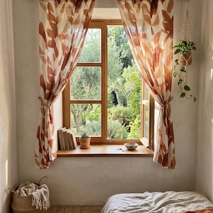 May include: A sunlit bedroom with a window featuring leaf-patterned curtains in shades of brown and cream. A wooden window frame opens to a lush green garden. The room includes a bed with neutral bedding, a woven basket, and a hanging plant.