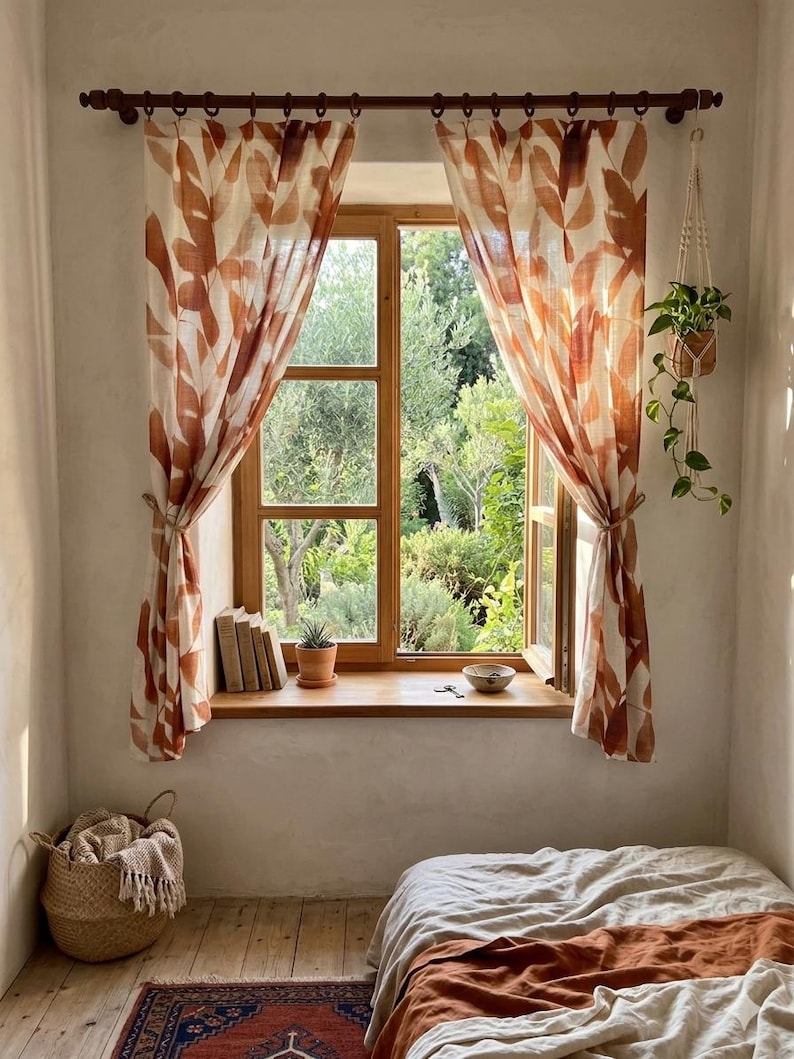 May include: A sunlit bedroom with a window featuring leaf-patterned curtains in shades of brown and cream. A wooden window frame opens to a lush green garden. The room includes a bed with neutral bedding, a woven basket, and a hanging plant.