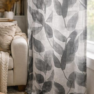 May include: A pair of sheer curtains with a large-scale leaf pattern in shades of gray and white. The curtains are hanging in front of a window, with a glimpse of a white sofa and a bookshelf in the background. The fabric appears to be linen.