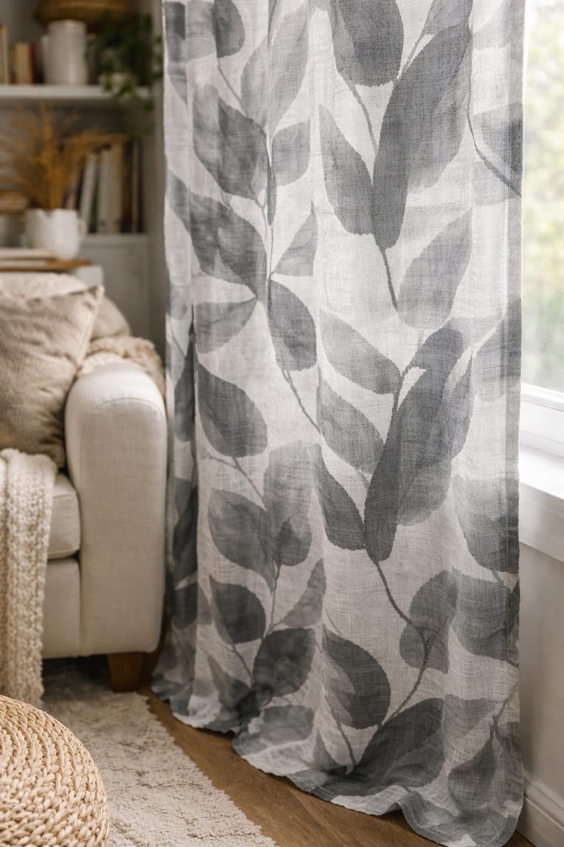 May include: A pair of sheer curtains with a large-scale leaf pattern in shades of gray and white. The curtains are hanging in front of a window, with a glimpse of a white sofa and a bookshelf in the background. The fabric appears to be linen.