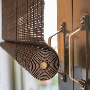 Könnte beinhalten: Aufgerollter brauner Bambus-Fensterrollo. Der Rollo besteht aus dünnen, rechteckigen Bambusstücken, die miteinander verwoben sind. Der Rollo ist teilweise aufgerollt und zeigt den Holzdübel in der Mitte. Er befindet sich neben einer dunkelbraunen Tür mit Metallgriffen.