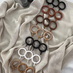 May include: An assortment of curtain rings in various colors and finishes, including black, white, and several shades of wood. The rings are arranged on a textured, neutral-toned fabric, with a few gray candles in the background.