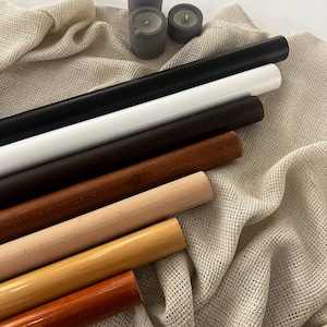 Op de afbeelding: Een verzameling houten deuvels in verschillende tinten bruin, beige en natuurlijk hout, samen met zwart en wit geschilderde deuvels. Drie kleine grijze betonnen kaarshouders met brandende kaarsen liggen op een beige stoffen achtergrond.