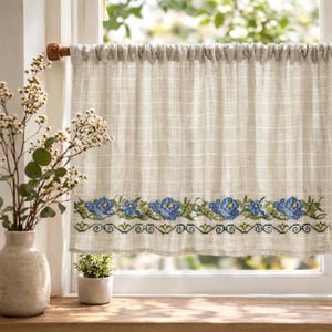 Könnte beinhalten: Ein beiger Leinen-Fenstervorhang mit einem Gittermuster und einem floralen Kreuzstich-Bordüren in Blau und Grün. Der Vorhang hängt an einer Holzstange vor einem Fenster.