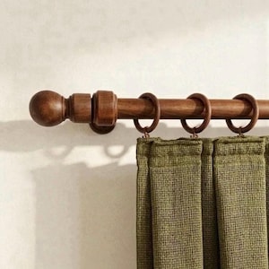 May include: A wooden curtain rod with a ball finial and metal rings. Olive green textured curtains hang below. The rod and finial are a warm brown color, creating a rustic and natural aesthetic.
