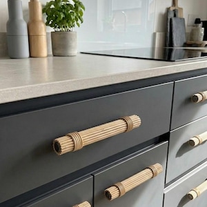 Op de afbeelding: Een keukenkast met donkergrijze deuren en natuurlijke rotan handgrepen. De handgrepen zijn cilindrisch met een geribbelde textuur. Een aanrechtblad bevat een potplant, zout- en pepermolens.