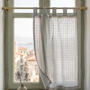 Puede incluir: Una cortina transparente de color gris claro con un patrón de cuadrícula cuelga frente a una ventana. La cortina está suspendida de una barra dorada. Un jarrón de lavanda se encuentra en el alféizar de la ventana, con vistas a un pueblo al fondo.