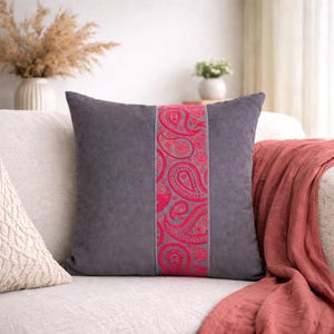 May include: A square grey cushion featuring a central band of bright pink paisley pattern. The cushion sits on a cream sofa, with a red-orange throw draped over the arm. A vase of dried flowers is in the background.