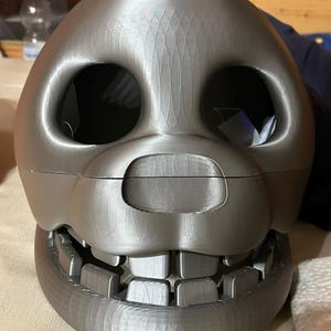 May include: A gray, 3D-printed skull mask with large eye sockets and a wide, toothy grin. The mask has a textured surface and appears to be made of plastic. The design is reminiscent of a cartoon character or a costume prop.