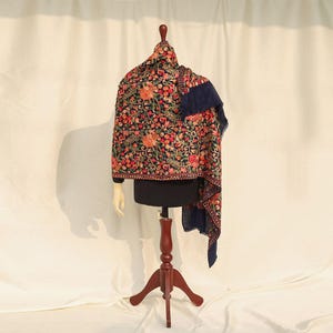May include: A dark navy blue shawl featuring dense, multi-colored floral embroidery in shades of orange, red, pink, and green. The intricate needlework covers the fabric, showcasing detailed botanical patterns across the entire surface area.