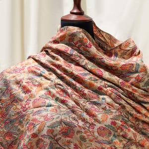May include: A beige shawl featuring intricate, multi-colored floral embroidery in shades of pink, orange, green, and blue. The fabric is draped over a mannequin, showcasing a dense, continuous pattern of flowers, leaves, and vines across the surface.