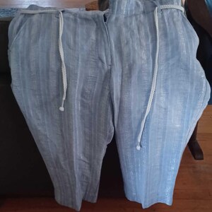 Puede incluir: Pantalones a rayas azul claro con cord&oacute;n blanco. Los pantalones tienen un patr&oacute;n de rayas verticales y un ajuste relajado. La tela parece tener un brillo sutil. Los pantalones est&aacute;n doblados y expuestos sobre una superficie oscura.