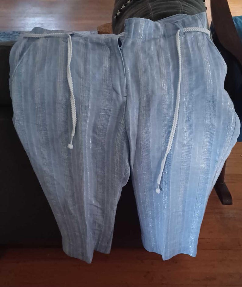 Puede incluir: Pantalones a rayas azul claro con cord&oacute;n blanco. Los pantalones tienen un patr&oacute;n de rayas verticales y un ajuste relajado. La tela parece tener un brillo sutil. Los pantalones est&aacute;n doblados y expuestos sobre una superficie oscura.