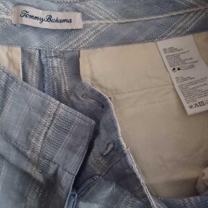 Puede incluir: Primer plano de unos pantalones cortos azul claro con un patr&oacute;n texturizado. Los pantalones cortos tienen cierre de bot&oacute;n y un bolsillo interior blanco. La etiqueta de la marca Tommy Bahama es visible.