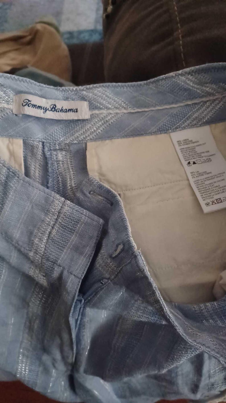 Puede incluir: Primer plano de unos pantalones cortos azul claro con un patr&oacute;n texturizado. Los pantalones cortos tienen cierre de bot&oacute;n y un bolsillo interior blanco. La etiqueta de la marca Tommy Bahama es visible.