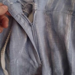 Puede incluir: Primer plano de tela azul claro con rayas plateadas verticales. La tela tiene una cremallera y un cord&oacute;n blanco. La imagen muestra una parte de la prenda, posiblemente pantalones o una falda.