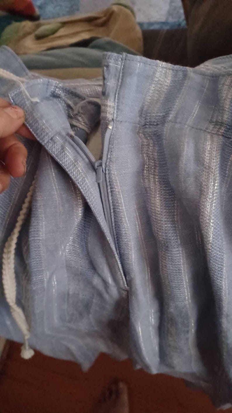 Puede incluir: Primer plano de tela azul claro con rayas plateadas verticales. La tela tiene una cremallera y un cord&oacute;n blanco. La imagen muestra una parte de la prenda, posiblemente pantalones o una falda.