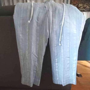 Puede incluir: Pantalones azul claro con rayas verticales y cintura con cord&oacute;n. La tela ligera parece ligeramente brillante. Los pantalones est&aacute;n colgados sobre una superficie marr&oacute;n oscuro.