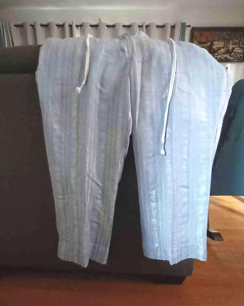 Puede incluir: Pantalones azul claro con rayas verticales y cintura con cord&oacute;n. La tela ligera parece ligeramente brillante. Los pantalones est&aacute;n colgados sobre una superficie marr&oacute;n oscuro.