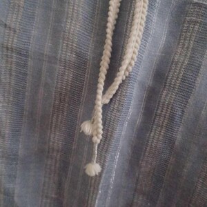 Puede incluir: Primer plano de una tela azul claro con rayas verticales y un cord&oacute;n blanco. El cord&oacute;n es una trenza con peque&ntilde;as borlas decorativas. La tela tiene un brillo sutil.