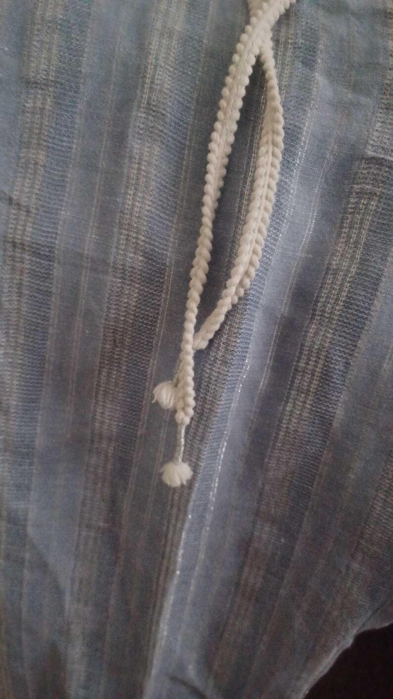 Puede incluir: Primer plano de una tela azul claro con rayas verticales y un cord&oacute;n blanco. El cord&oacute;n es una trenza con peque&ntilde;as borlas decorativas. La tela tiene un brillo sutil.