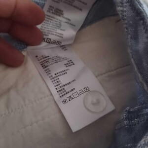 Puede incluir: Primer plano del interior de unos pantalones vaqueros azul claro. Se ven dos etiquetas blancas con texto y s&iacute;mbolos de cuidado. Tambi&eacute;n se ve un bot&oacute;n blanco. La tela es de color azul claro.