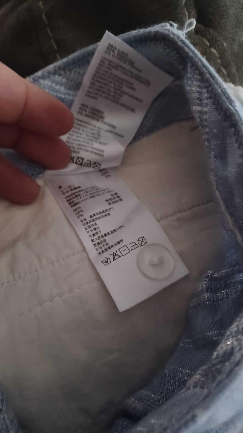 Puede incluir: Primer plano del interior de unos pantalones vaqueros azul claro. Se ven dos etiquetas blancas con texto y s&iacute;mbolos de cuidado. Tambi&eacute;n se ve un bot&oacute;n blanco. La tela es de color azul claro.