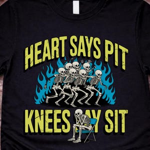 Puede incluir: Camiseta negra con un diseño de esqueleto humorístico. La camiseta presenta el texto "HEART SAYS PIT KNEES SAY SIT" en amarillo, con un grupo de esqueletos bailando y un esqueleto sentado.