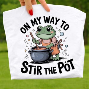 Puede incluir: Camiseta blanca con un dibujo de una rana de dibujos animados removiendo una olla, con un delantal. El texto "ON MY WAY TO STIR THE PÖT" está impreso en negro. La rana es verde con ojos grandes y sostiene una cuchara de madera.