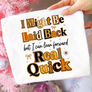 以下が含まれることがあります： 「I Might Be Laid Back but I can lean forward Real Quick」のテキストがプリントされた白い正方形の布。テキストは、ヒョウ柄やオレンジ色など、さまざまなフォントと色を使用しています。小さなリボンとスターバーストがテキストを飾っています。