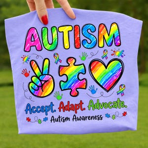 May include: A light purple t-shirt with the word "AUTISM" in rainbow colors. The shirt features a peace sign, puzzle piece, and heart, all in rainbow colors, along with the words "Accept. Adapt. Advocate." and "Autism Awareness."