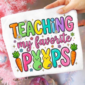 May include: White t-shirt with the text "TEACHING my favorite PEEPS" in colorful bubble letters. The design includes cartoon bunnies, carrots, and flowers. The shirt is held by a hand, suggesting it's a wearable item.