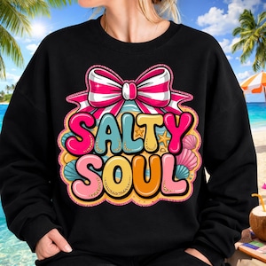 Può includere: Felpa nera con una grafica colorata che recita "Salty Soul". Il design include un fiocco rosa e bianco, conchiglie e le parole in colori vivaci. Lo sfondo mostra una scena di spiaggia.