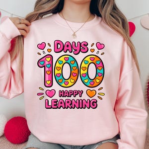 Peut inclure: Un sweat-shirt rose clair avec un graphisme coloré célébrant 100 jours d'école. Le motif présente les mots "DAYS", "100" et "HAPPY LEARNING" dans une police ludique, décorés de cœurs et d'étoiles.