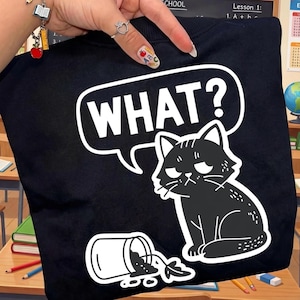 Puede incluir: Sudadera negra con un gráfico blanco de un gato gruñón y la palabra "WHAT?" en un bocadillo. También hay un recipiente derramado con hojas. La sudadera es sostenida por una persona.
