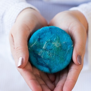 May include: A round, blue and green soap bar held in cupped hands. The soap is made of natural fibres and has a textured surface.