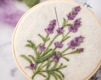 May include: A close-up of a needle felted lavender flower design in a wooden embroidery hoop. The hoop is on a white marble surface. There are small piles of purple and green wool fibers nearby.