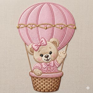 May include: Embroidered design of a teddy bear in a pink hot air balloon. The bear wears a pink bow and dress, waving from a woven basket. The balloon is pink with gold accents, set against a neutral fabric background.