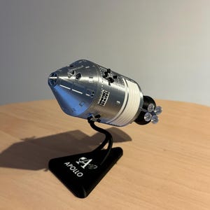 Pode incluir: Um modelo de nave espacial Apollo prateado e branco com as palavras "UNITED STATES" impressas na lateral. O modelo é montado em um suporte preto que diz "APOLLO". A nave espacial tem uma frente cônica e um corpo cilíndrico.
