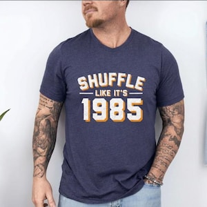 May include: A navy blue t-shirt featuring the text "SHUFFLE LIKE IT'S 1985" in white and orange. The t-shirt is a crew neck style. A denim jacket hangs on the wall in the background. The person is wearing blue jeans.