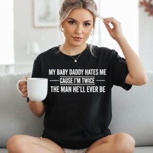 May include: Black t-shirt with the text "MY BABY DADDY HATES ME - CAUSE I'M TWICE THE MAN HE'LL EVER BE" in white. The shirt is a casual style. A white mug is held in the left hand.