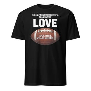 May include: Black t-shirt featuring the text "THE ONLY THING MORE POWERFUL THAN HATE IS LOVE" above a brown American football with the text "TOGETHER, WE ARE AMERICA".