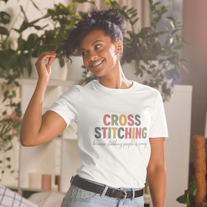May include: White t-shirt featuring the words "CROSS STITCHING" in various colours, with the phrase "because stabbing people is wrong" underneath. The person wearing the shirt is also wearing blue jeans and a black belt.