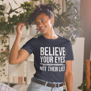 Peut inclure: Un t-shirt bleu marine avec le texte "BELIEVE YOUR EYES NOT THEIR LIES" en lettres blanches. Le t-shirt est porté par une personne aux cheveux foncés et est associé à un jean bleu et une ceinture noire.