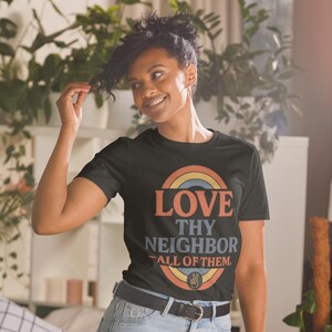 Puede incluir: Camiseta negra con un diseño de arcoíris y las palabras "LOVE THY NEIGHBOR - ALL OF THEM". El diseño incluye un símbolo de la paz. La camiseta está hecha de un material suave.