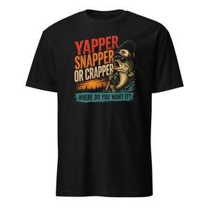 May include: A black t-shirt featuring a graphic of a fish wearing sunglasses and a cap while holding a fishing rod. The text reads YAPPER SNAPPER OR CRAPPER and WHERE DO YOU WANT IT? The design includes a sunset over a forest silhouette.
